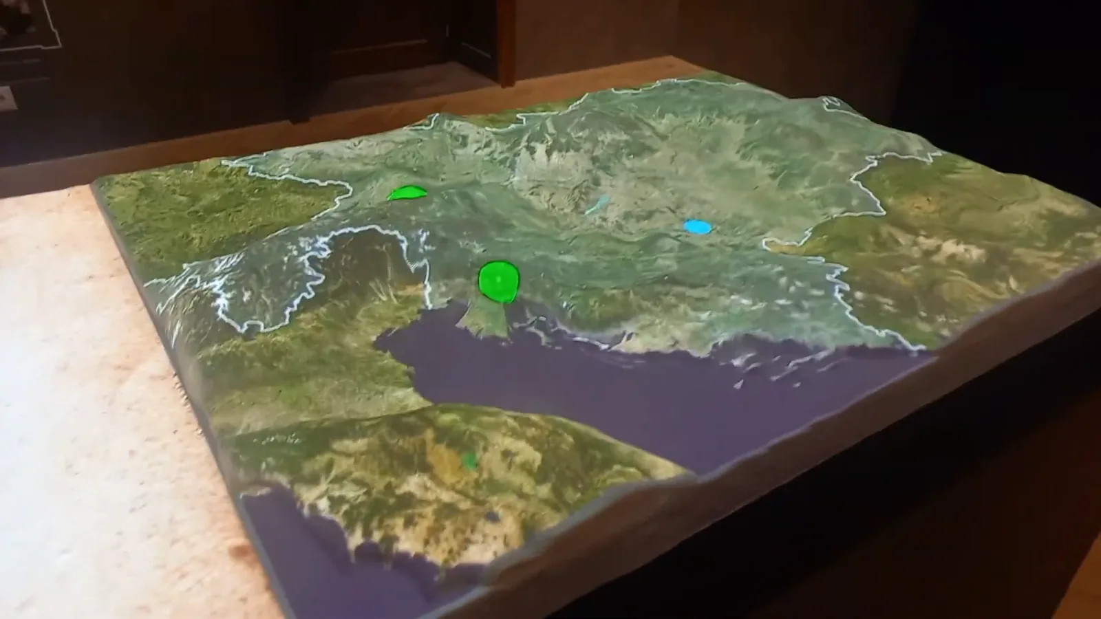 The relief model in the exhibition room, with borders traced in light across the terrain and points of interest marked in green and blue.